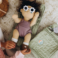 Dinkum Dolls doll in a mauve romper and flower sunglasses rests on a sage quilted changing mat while a child adjusts her shoes.
