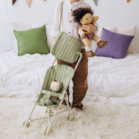 Child holds a doll beside a stroller with a sage green quilted changing bag and doll seat, showcasing the Dinkum Dolls Carrie Convertible Changing Set.