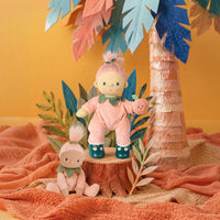 A styled product display featuring two plush dolls in pink outfits, one standing on a wooden stump and one sitting on a peach-colored blanket, with a paper palm tree and tropical leaves in the background against a warm yellow wall.