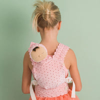 A young girl viewed from behind wearing a pink polka-dot baby carrier wrap with a matching bonnet featuring a sleeping doll face, set against a solid pale green background.