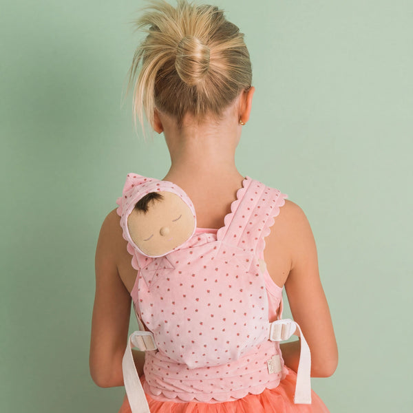 A young girl viewed from behind wearing a pink polka-dot baby carrier wrap with a matching bonnet featuring a sleeping doll face, set against a solid pale green background.