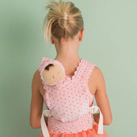 A young girl viewed from behind wearing a pink polka-dot baby carrier wrap over her shoulders, with a matching bonnet and a peach tulle dress, set against a solid sage green background.