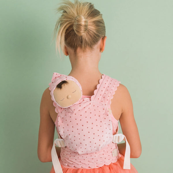 A young girl viewed from behind wearing a pink polka-dot baby carrier wrap over her shoulders, with a matching bonnet and a peach tulle dress, set against a solid sage green background.