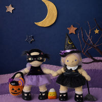 Two Dinkum Dolls wearing Halloween costumes, including a witch hat and black dress, alongside a cat costume with a purple tutu, pumpkin bucket, and crescent moon backdrop by Olli Ella