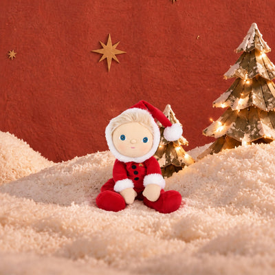 A small plush doll dressed in a red Santa-style outfit with white trim and a red hat sits on a cream textured surface against a warm red-orange backdrop, with decorative gold stars and lit Christmas trees behind it.