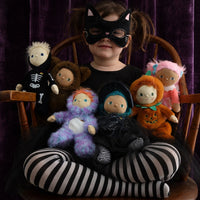Child in a cat mask surrounded by colorful Dinky Dinkum dolls in Halloween costumes, including a pink-haired doll, against a dark velvet backdrop.