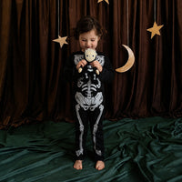 A child in a skeleton costume holds a small Scully Skeleton doll against a dark curtain with hanging stars and a crescent moon from Dinkum Dolls by Olli Ella