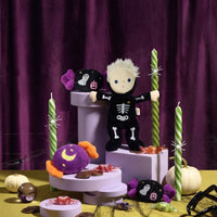 Dinky Dinkum Scully Skeleton doll in a black bone-print outfit with blond hair, surrounded by striped candles, pumpkins and sweets in a spooky display by Dinkum Dolls by Olli Ella