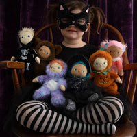 A child in a black cat mask sits with Dinky Dinkum dolls in Halloween costumes, including Wesley Werewolf from Dinkum Dolls by Olli Ella.