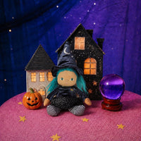 Dinky Dinkum Winnie Witch doll with green hair and a black witch hat sits beside a pumpkin and crystal ball in a spooky Halloween village display by Dinkum Dolls by Olli Ella.