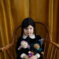 Child in a black dress holds two Dinky Dinkum dolls, including green-haired Winnie Witch, seated in a wooden chair before a golden velvet curtain.