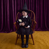 Child in a black witch costume sits in a wooden chair while holding the black Dozy Dinkum Willa Witch doll from Dinkum Dolls by Olli Ella.
