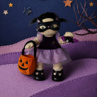 Dream Dinkum doll Mabel Moon dressed as a witch with a cat mask holds a pumpkin trick or treat basket and a Halloween lollipop on a spooky quilted backdrop