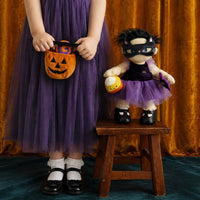Child in a purple witch costume holds a pumpkin basket beside Mabel Moon doll in a purple tutu, black mask and boots on a wooden stool by Dinkum Dolls by Olli Ella