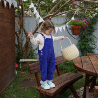Child holding a woven straw gathering basket while standing on a garden bench outdoors, with trees, bunting, and flowers in the background by Olli Ella Kids Rattan