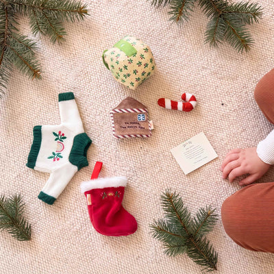 Dinkum Doll Christmas accessories laid on a cream knit rug with pine sprigs: a knitted holly jumper, red stocking, plush gift box, candy cane, letter to Santa and a note card.
