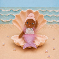 A small mermaid doll with brown skin and blue eyes sits inside a large beige seashell on a sandy surface, with a blue ocean backdrop and scattered pearl-like beads.
