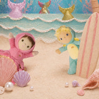 Two small plush mermaid dolls on a sandy beach scene with shells, pearls, a surfboard, and mermaid tail decorations in the background.