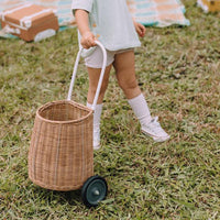 A child pulls a natural rattan wheeled basket across the grass, showing the Olli Ella Kids Rattan Original Luggy for outdoor play and storage.
