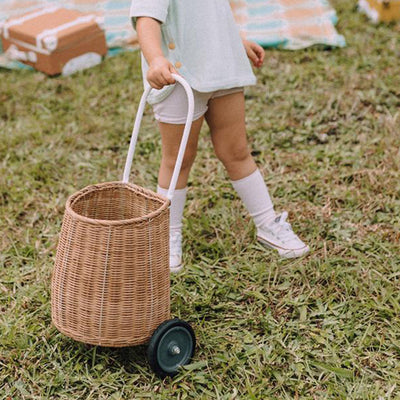 A child pulls a natural rattan wheeled basket across the grass, showing the Olli Ella Kids Rattan Original Luggy for outdoor play and storage.