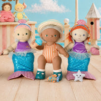 Three mermaid-themed dolls seated on a wooden deck with a beach and sky backdrop. The center doll wears an orange-and-white striped swimsuit and white hat, while the two side dolls wear purple tops and blue mermaid tails with pink fins.
