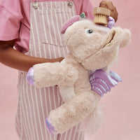 Dinkum Pets plush pink unicorn with lavender wings and hooves, held by a child in a striped apron while being gently brushed