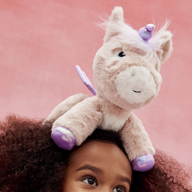 Pink plush Dinkum Unicorn Shimmer with a purple horn and hooves perched on a childs head against a soft pink background from Dinkum Pets