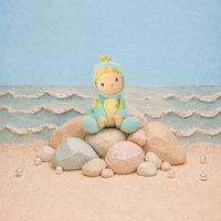 A plush doll with a yellow face and blonde hair sits on a stack of rocks on a sandy beach set. The doll is wearing a light blue hooded outfit with a yellow belly and small yellow accents.