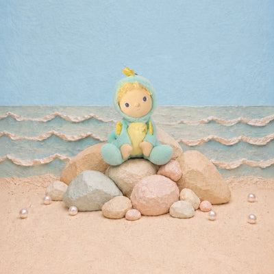 A plush doll with a yellow face and blonde hair sits on a stack of rocks on a sandy beach set. The doll is wearing a light blue hooded outfit with a yellow belly and small yellow accents.