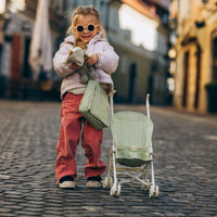 Child wearing sunglasses holds a doll beside a sage green stroller and matching changing bag on a cobblestone street, showcasing the Dinkum Dolls Carrie set.