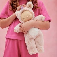 Child in pink clothing hugs a soft pink Cozy Dinkums unicorn plush with a purple horn, floppy ears, and fluffy wings from Dozy Dinkums.