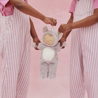 Two children in pink striped overalls hold a soft lavender unicorn plush doll with a cream face and white belly against a pink background from Dozy Dinkums