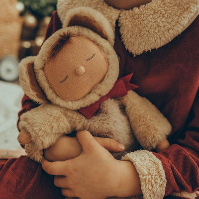 Cozy Dinkums Teddy Crumble plush doll with a sleeping face and bear hood, held by a child in a red outfit with a soft cream fleece collar and cuffs
