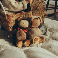 Two brown Cozy Dinkums plush dolls with round ears and red scarves sit together on cream bedding beside a woven wicker basket in a cozy nursery setting
