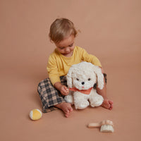 Child cuddling a fluffy white Dinkum Dogs toy puppy wearing a red bandana, with a grooming brush and toy ball nearby for imaginative play.