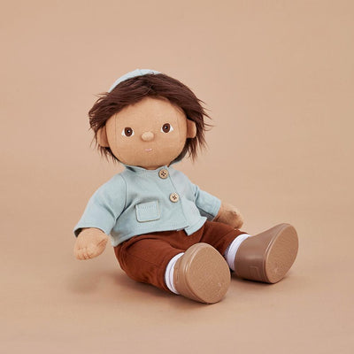 Dinkum Dolls doll wearing a pale blue hooded jacket and brown pants with white socks and brown shoes, seated against a warm neutral background.