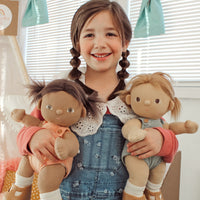 Smiling child in denim overalls cuddles two soft Dinkum Dolls, one with dark skin and brown hair and one with light skin and blonde hair indoors
