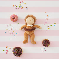 Dinky Dinkums Darcy Donut plush doll wearing a chocolate donut ring, surrounded by pink and chocolate donuts with colorful sprinkles on a striped pastel background