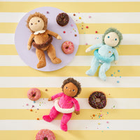 Dinky Dinkums plush dolls in pink, blue, and brown outfits surrounded by colorful sprinkled doughnuts and confetti on a striped yellow and white background