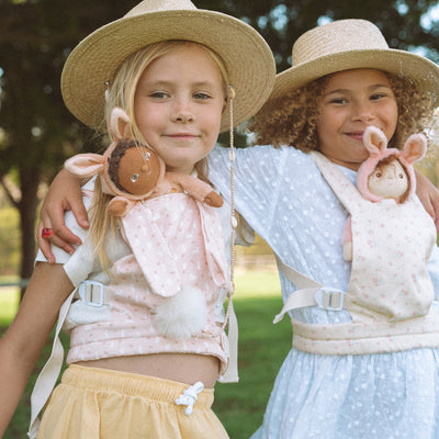 Two smiling children in sun hats play outdoors while holding soft Dinky Dinkums bunny dolls in apricot and cream pink outfits by Dinky Dinkums