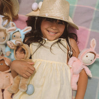 Smiling child in a straw hat cuddling several soft bunny dolls, including a pink plush bunny, against a pastel plaid background from Dinky Dinkums.