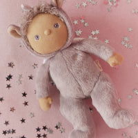 Dinky Dinkums Uma unicorn doll in a lilac purple plush hooded outfit, lying on a pink blanket scattered with silver and black star confetti.