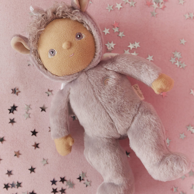 Dinky Dinkums Uma unicorn doll in a lilac purple plush hooded outfit, lying on a pink blanket scattered with silver and black star confetti.