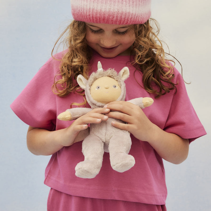 Child in a pink outfit holding a soft Dinky Dinkums Uma unicorn doll with a fluffy hood, ears, and horn from the Unicorn Collection