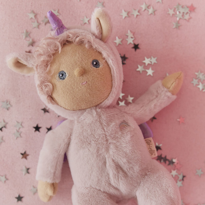 Dinky Dinkums Una unicorn doll in a soft pink plush outfit with a horn and wings, lying on a pink background scattered with colorful star confetti.