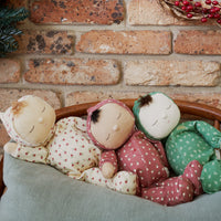 Custard Sweetheart Dozy Dinkum doll by Dinkum Dolls by Olli Ella rests with colorful floral dolls in a woven basket under a soft blanket against a brick wall