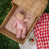 Dozy Dinkums Pip Strawberry doll in a pink strawberry print romper sleeping in a wicker basket beside a red gingham picnic blanket and fresh strawberry slice
