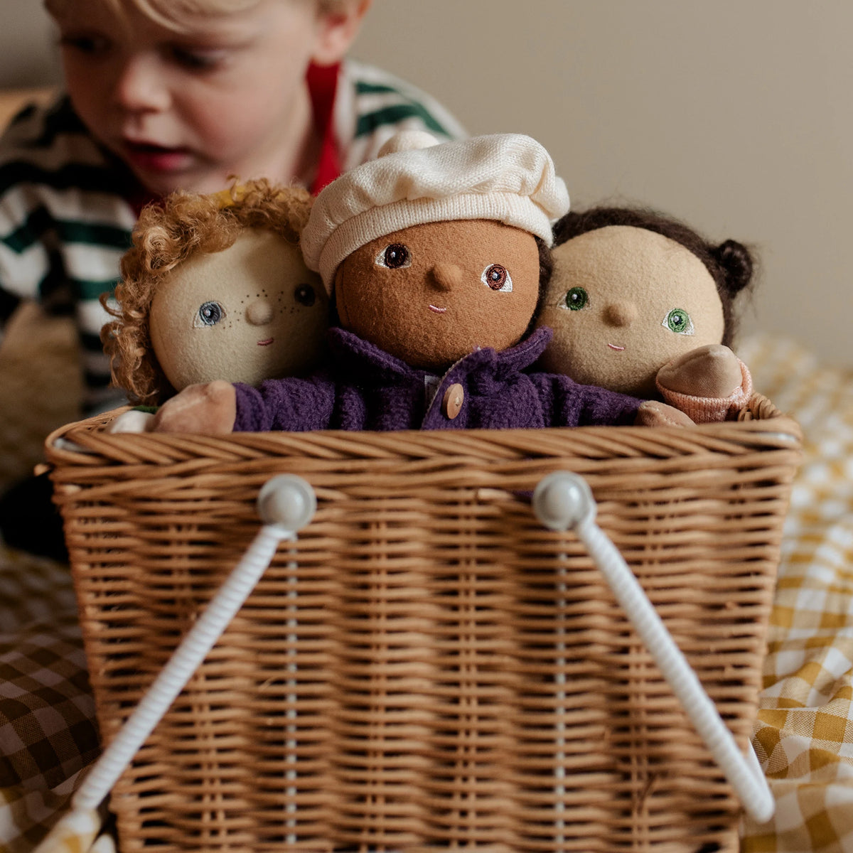 Three Dinky Dinkums dolls sit in a woven basket while a child looks on one doll wears a purple coat and cream cap