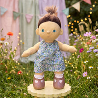 Doll in a floral dress standing on a wooden platform in a garden setting with flowers and lights.