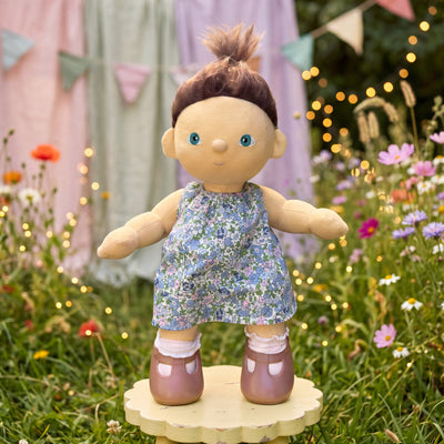 Doll in a floral dress standing on a wooden platform in a garden setting with flowers and lights.